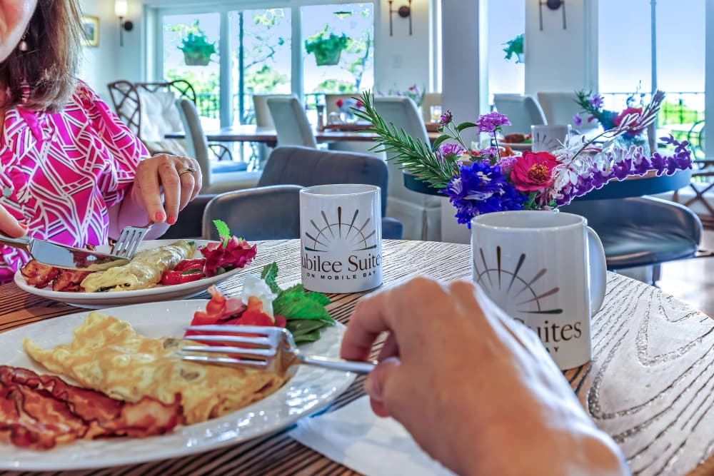 Some people sitting at a table eating a breakfast of omelets, bacon and fresh fruit, with cups of coffee, and fresh flowers.