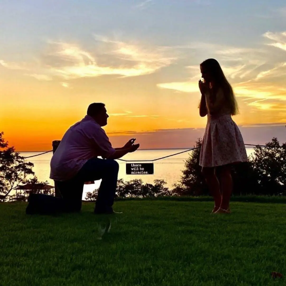 A man is proposing to a woman at sunset, while she appears surprised and emotional.