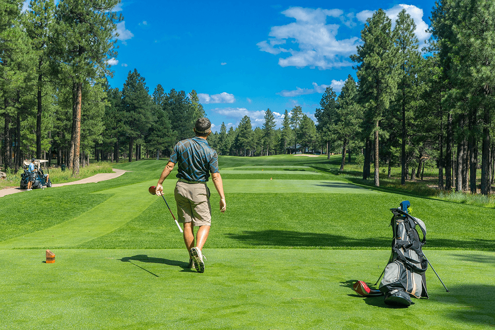 A golfer walks towards the fairway on a sunny day, with pine trees lining the back.