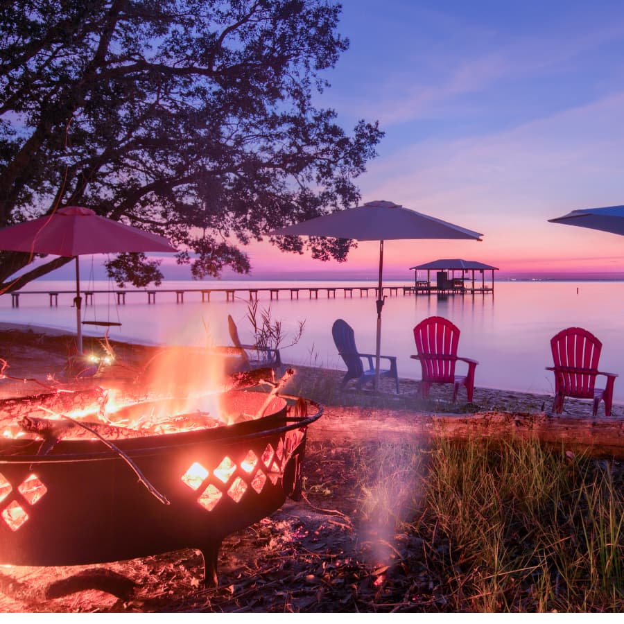 A cozy campfire burns in front of colorful chairs with a serene lakeside view at dusk.
