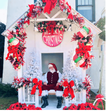 A smiling Santa sits in a festive gazebo decorated with red bows, ornaments, and white trees, under a "Merry Christmas" sign.