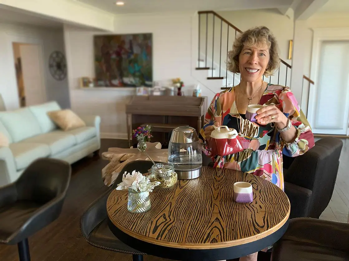 A smiling woman holds a teapot and cup while standing next to a table in a cozy living space.