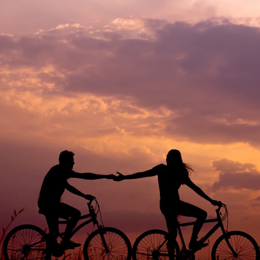 A man and woman riding bikes at sunset reaching out their hands to one another.