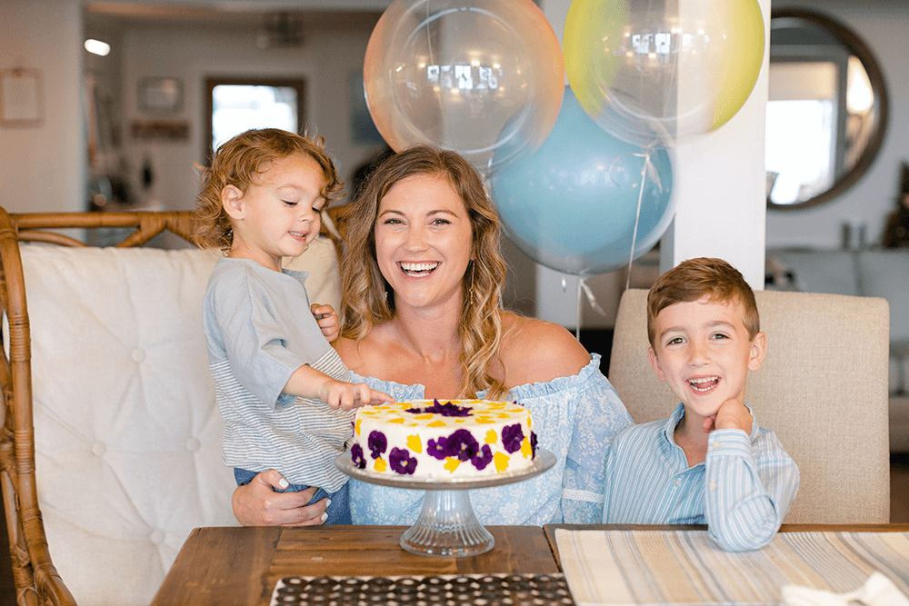 A smiling woman sits at a table with two children, celebrating with a cake and colorful balloons in the background.