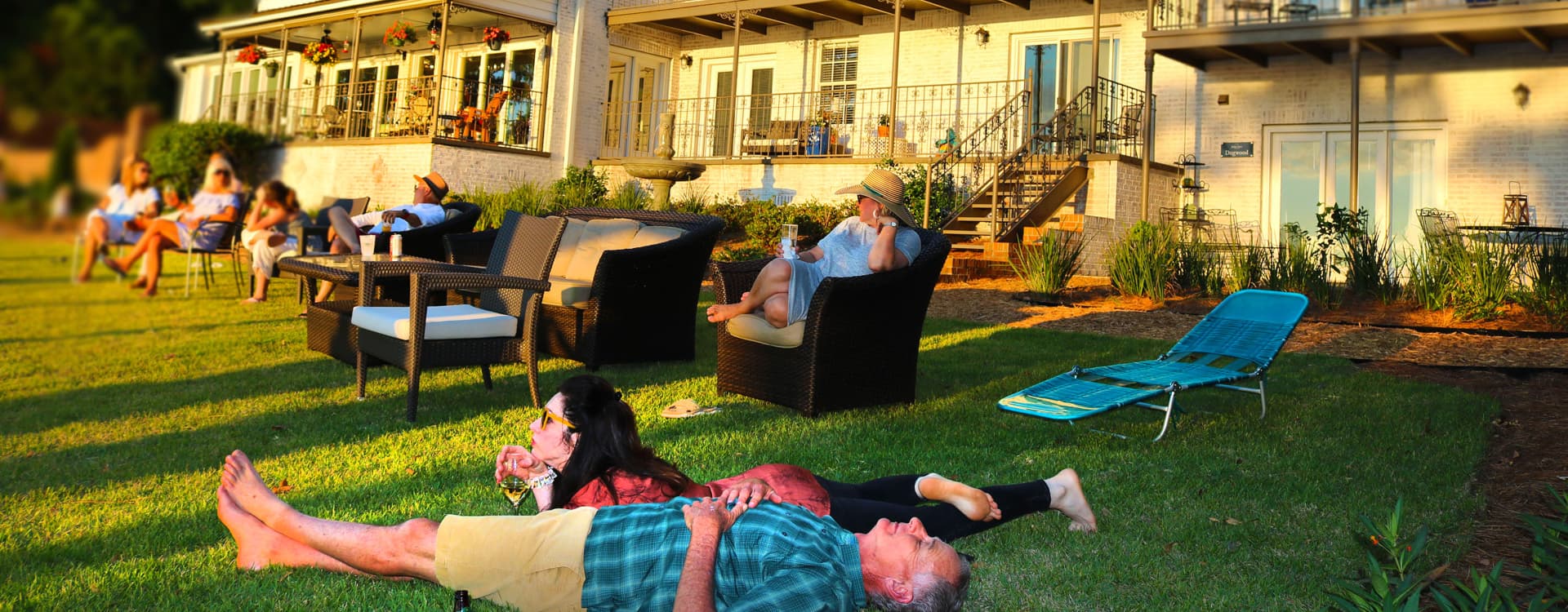 A group of people lounging around on the grass or in chairs, enjoying themselves in front of an expansive house.