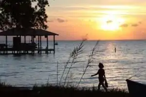 Silhouette of a child walking along a shoreline at sunset, with a pier in the background.