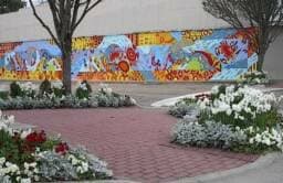 A vibrant mural is displayed on a wall behind a landscaped area filled with colorful flowers and a brick path.