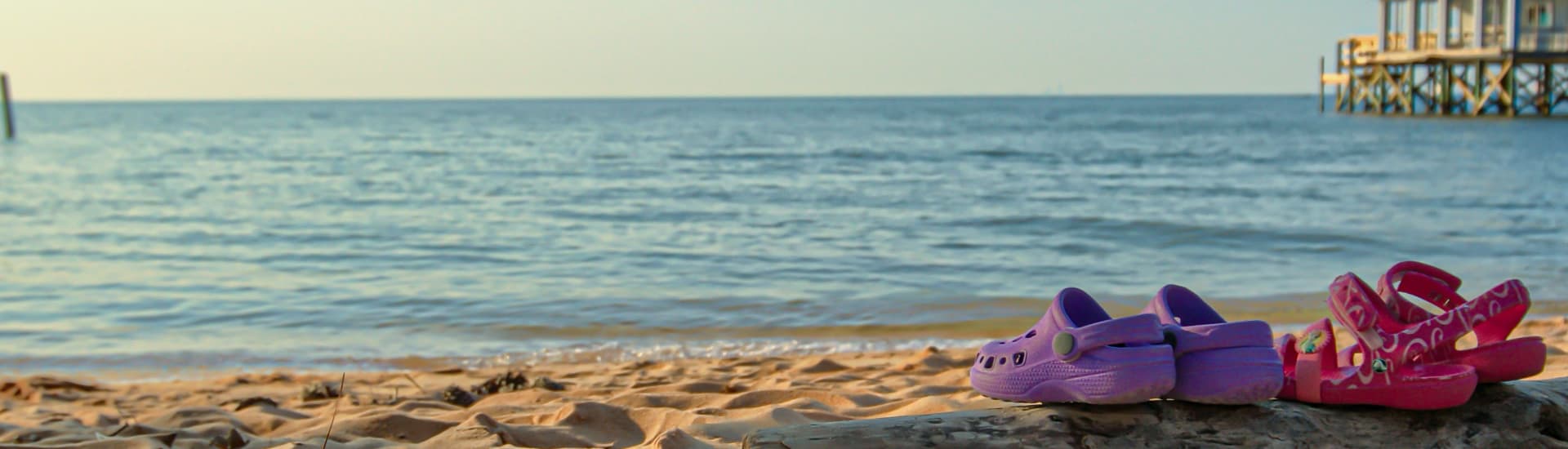 A pair of colorful Crocs sits on the sandy beach with calm ocean water in the background.
