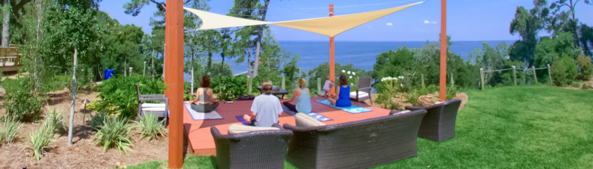 A group of people sitting on a covered deck with a shade awning meditating, overlooking the bright blue waters of Mobile Bay.