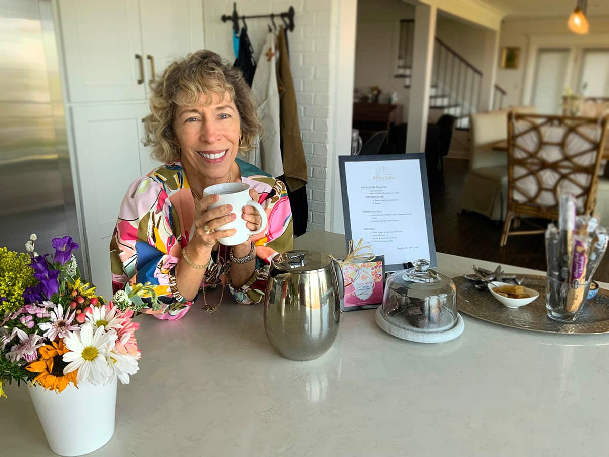 Smiling woman in a colorful blouse holding a cup, seated at a kitchen counter with flowers and a menu.