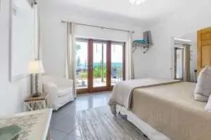 A bright and airy bedroom featuring a large bed, white armchair, and doors leading to an outdoor view. A bright and airy bedroom featuring a large bed, white armchair, and doors leading to an outdoor view.