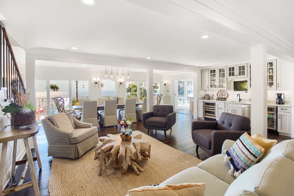 A large, open-plan living room with white walls, hardwood floors, and comfortable seating. Large windows offer stunning ocean views, and there's a dining table and a wet bar in the background.