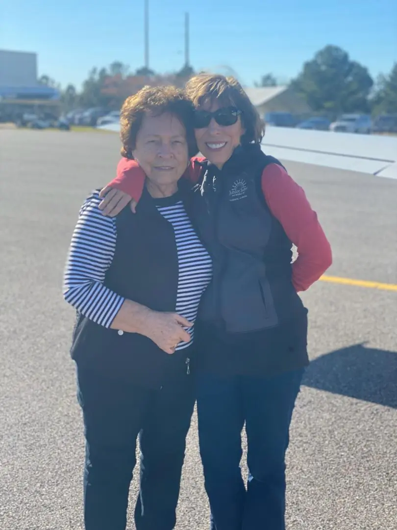 Two women, one in a striped shirt and the other in a jacket, embrace and smile at the camera outdoors.