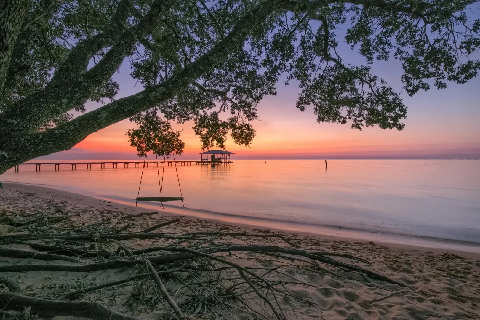 A serene beach at sunset with a swing, a wooden pier, and lush trees.
