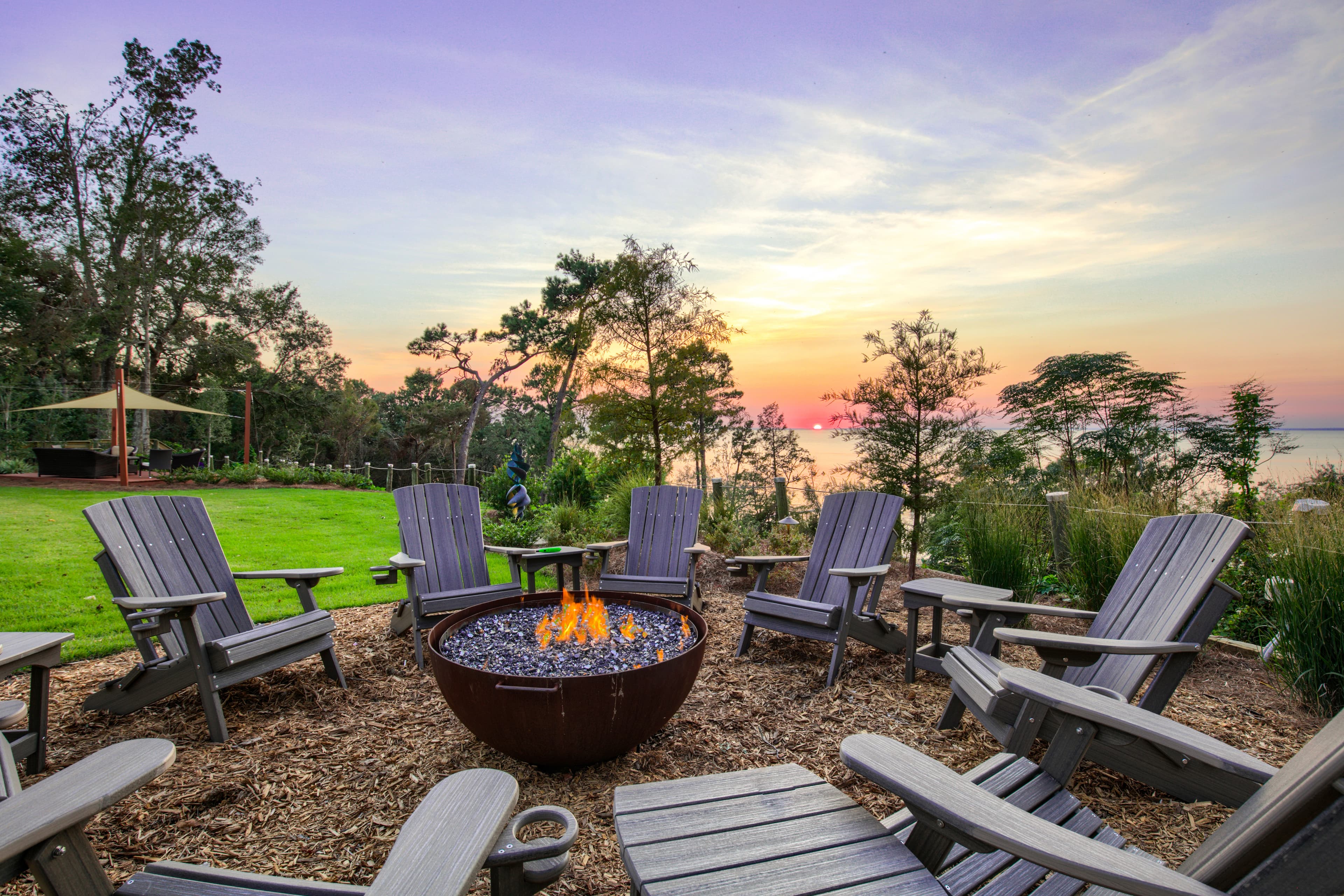 A scenic outdoor space with a fire pit surrounded by comfortable adirondack-style chairs. The sky is painted with vibrant hues of orange, pink, and purple as the sun sets over the water.