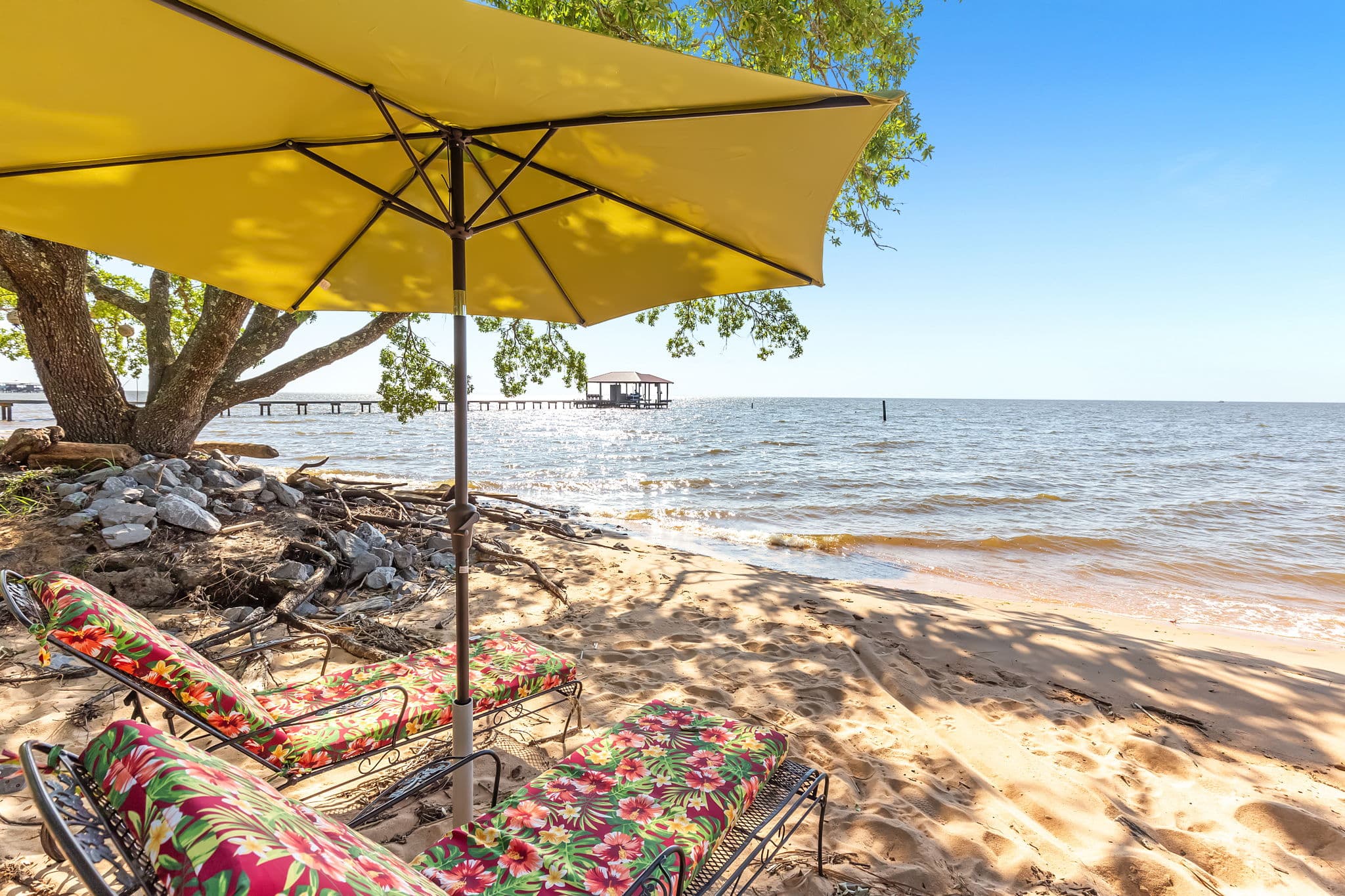 Two colorful beach chairs sit under a large yellow umbrella on a sandy beach on the bay.