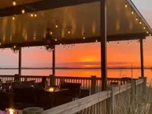 A cozy restaurant patio with string lights overlooks a vibrant orange sunset over the water.