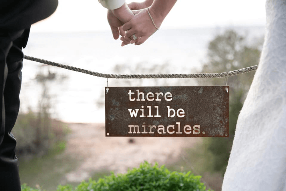 A close-up of two hands holding a sign that reads "there will be miracles," overlooking a scenic view.