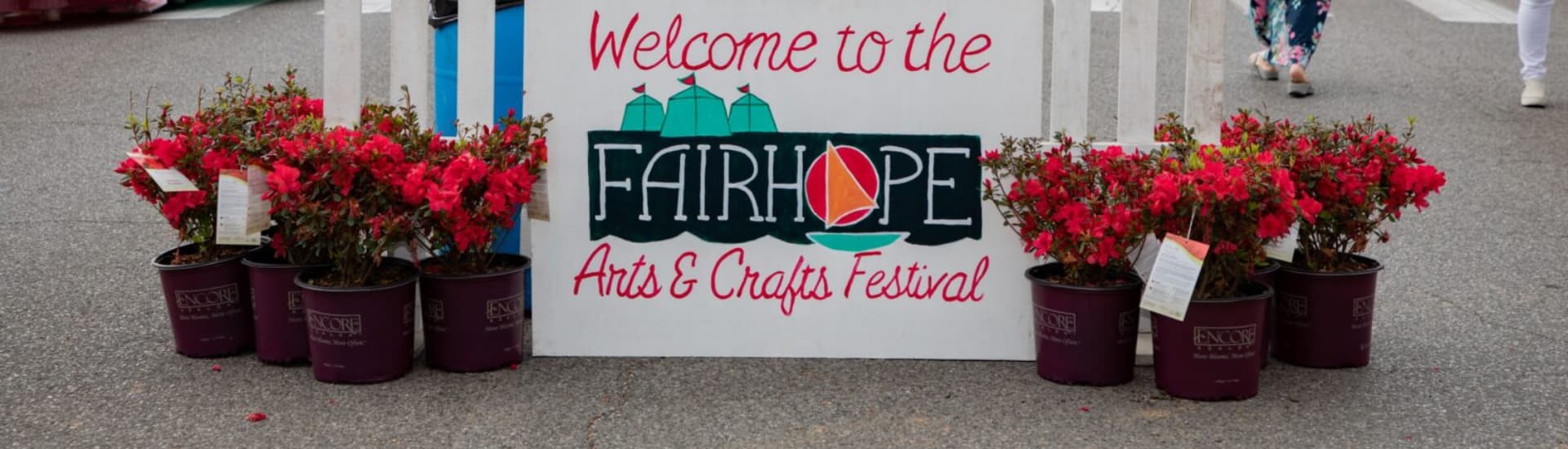 A sign welcoming visitors to the Fairhope Arts & Crafts Festival, with potted azaleas in front.