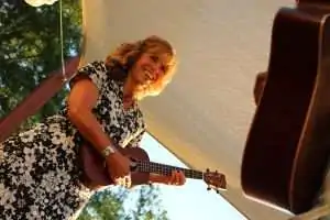 A smiling woman in a floral dress plays a ukulele under a canopy.