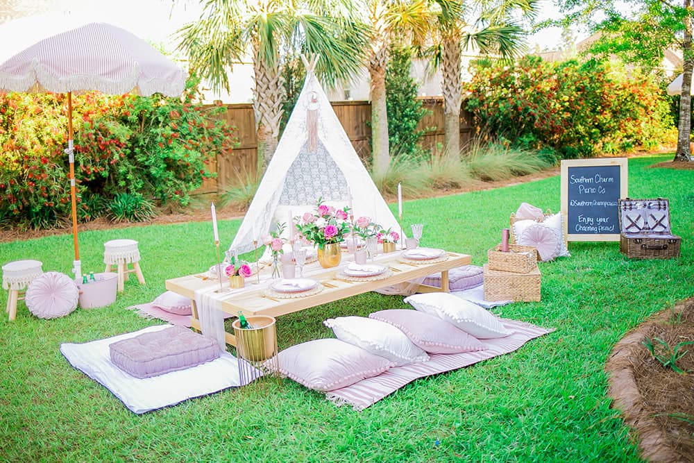 A green grassy area with an area set up with a low table beautiful set with dishes, glasses, tableware, and vases of fresh flowers, area rugs and cushions on the ground, an open tent, some stools and a shade umbrella, along with a picnic basket and a sandwich board that says Southern Charm Picnic Company.