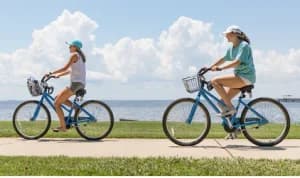 Two girls ride blue bicycles along a waterfront path under a bright, partly cloudy sky.