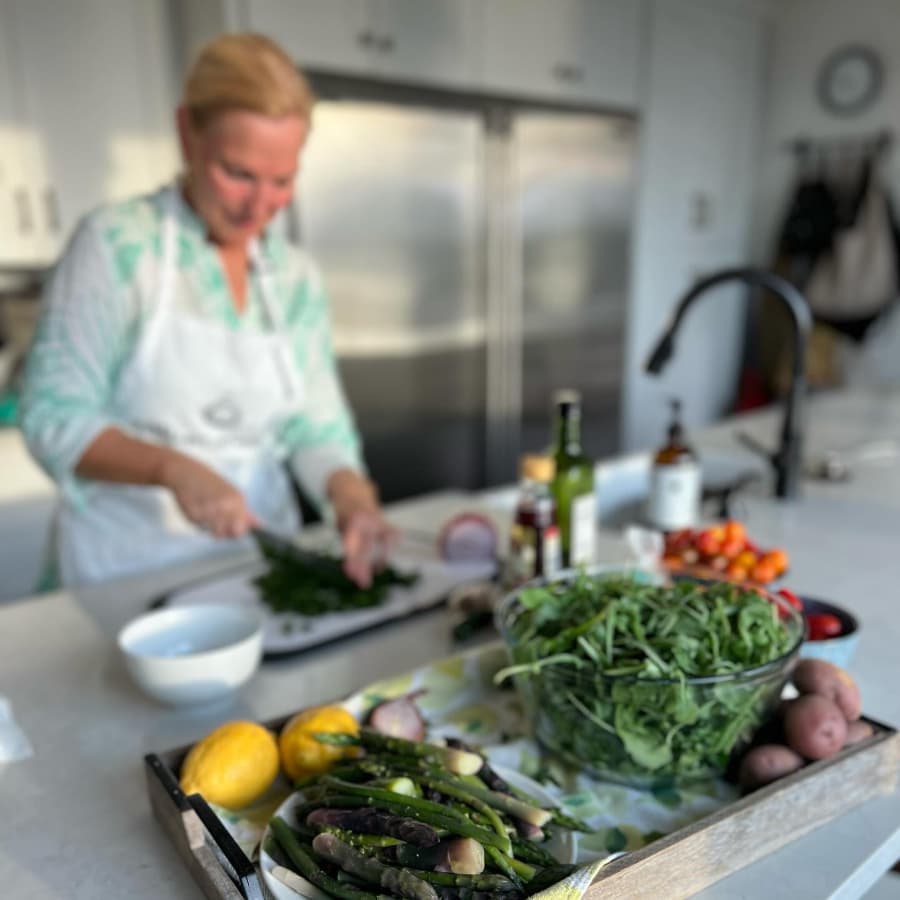 A wood tray with fresh vegetables and lemons, and a large bowl of greens for a salad, and a woman in the background chopping greens.