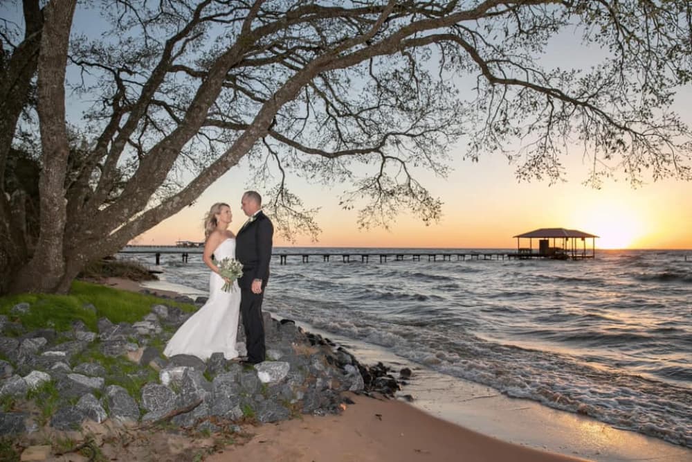 A bride and groom share a romantic moment by the water at sunset, surrounded by trees and stones.