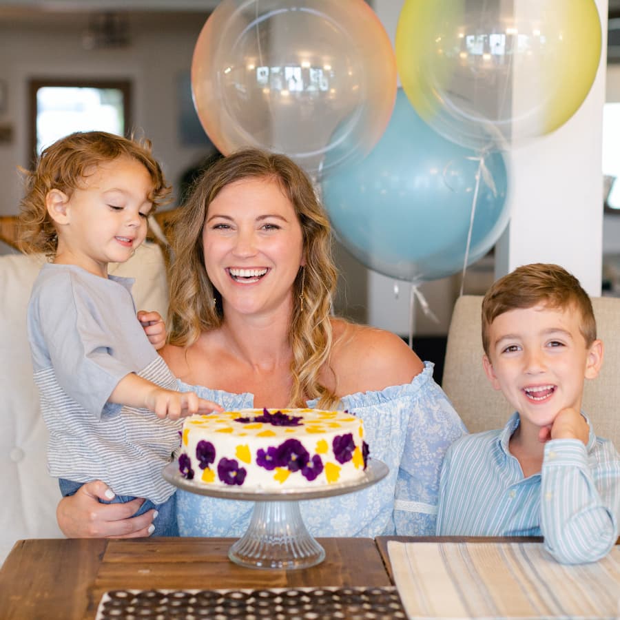 A smiling woman and 2 children in front of a decorated cake, and balloons behind them.